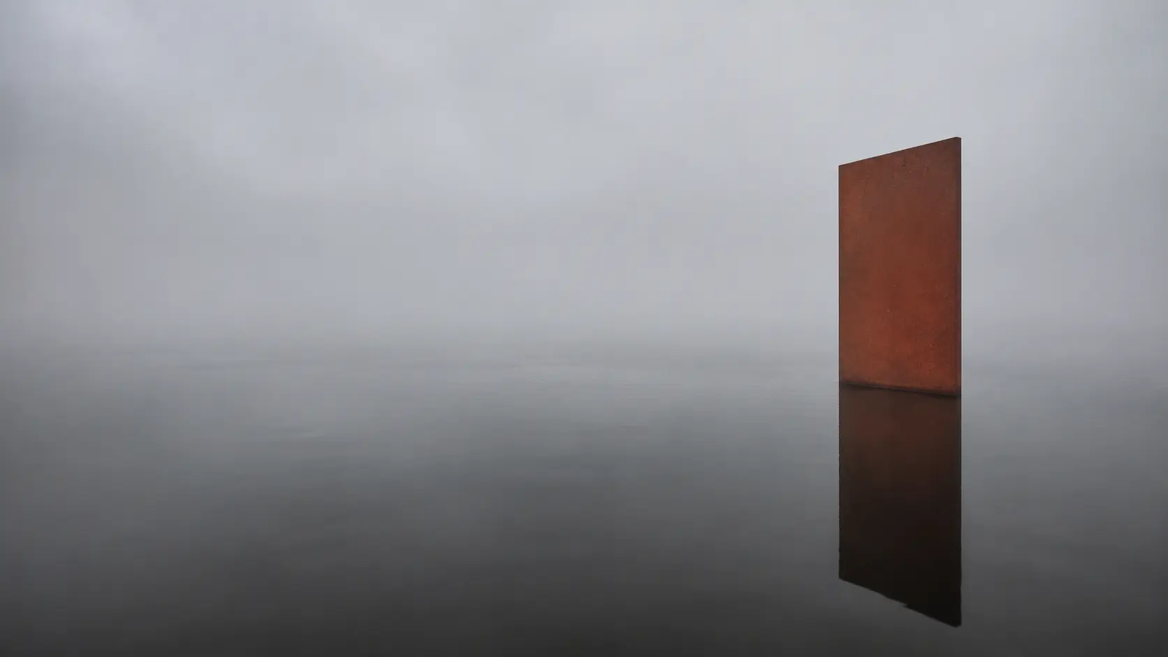 A plain rusted steel plate standing in still dark water and fog