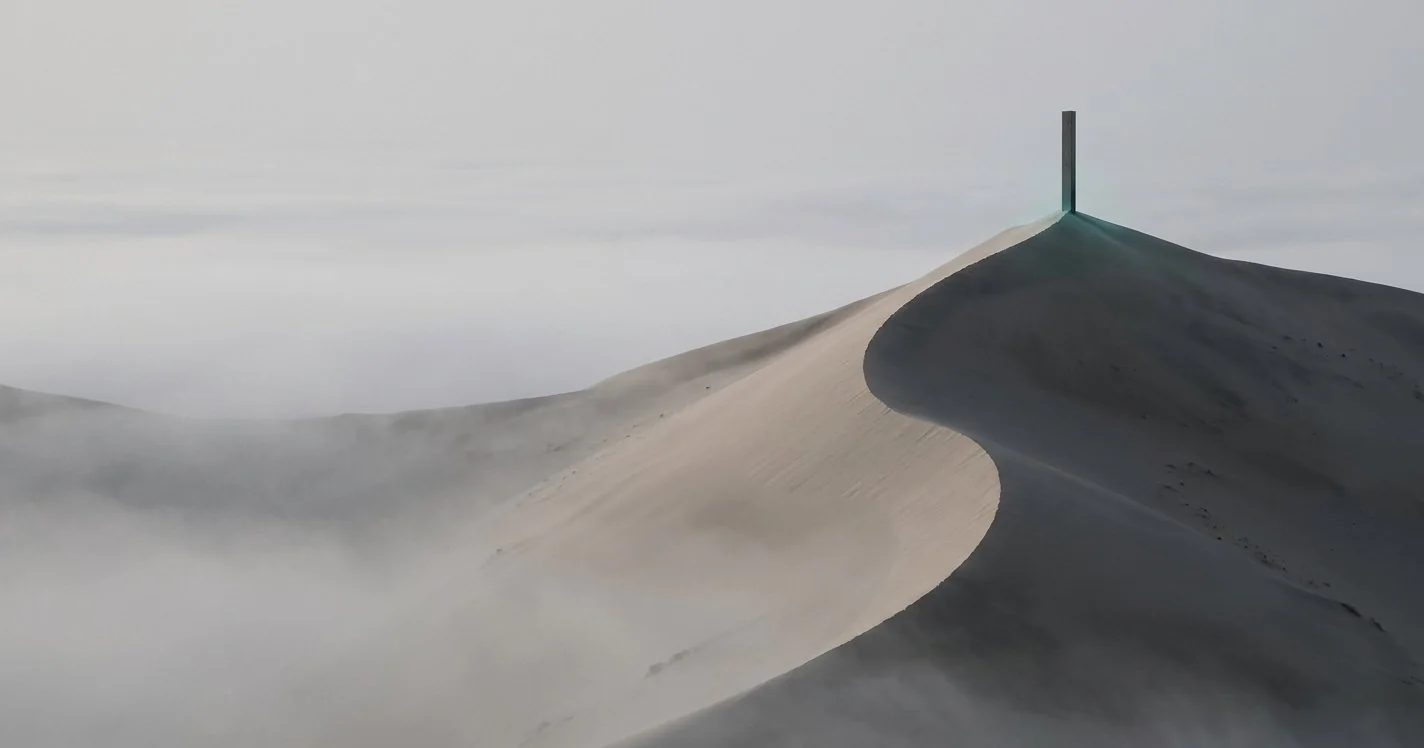 A slender dark monolith standing on a fog-covered Namib dune crest at dawn